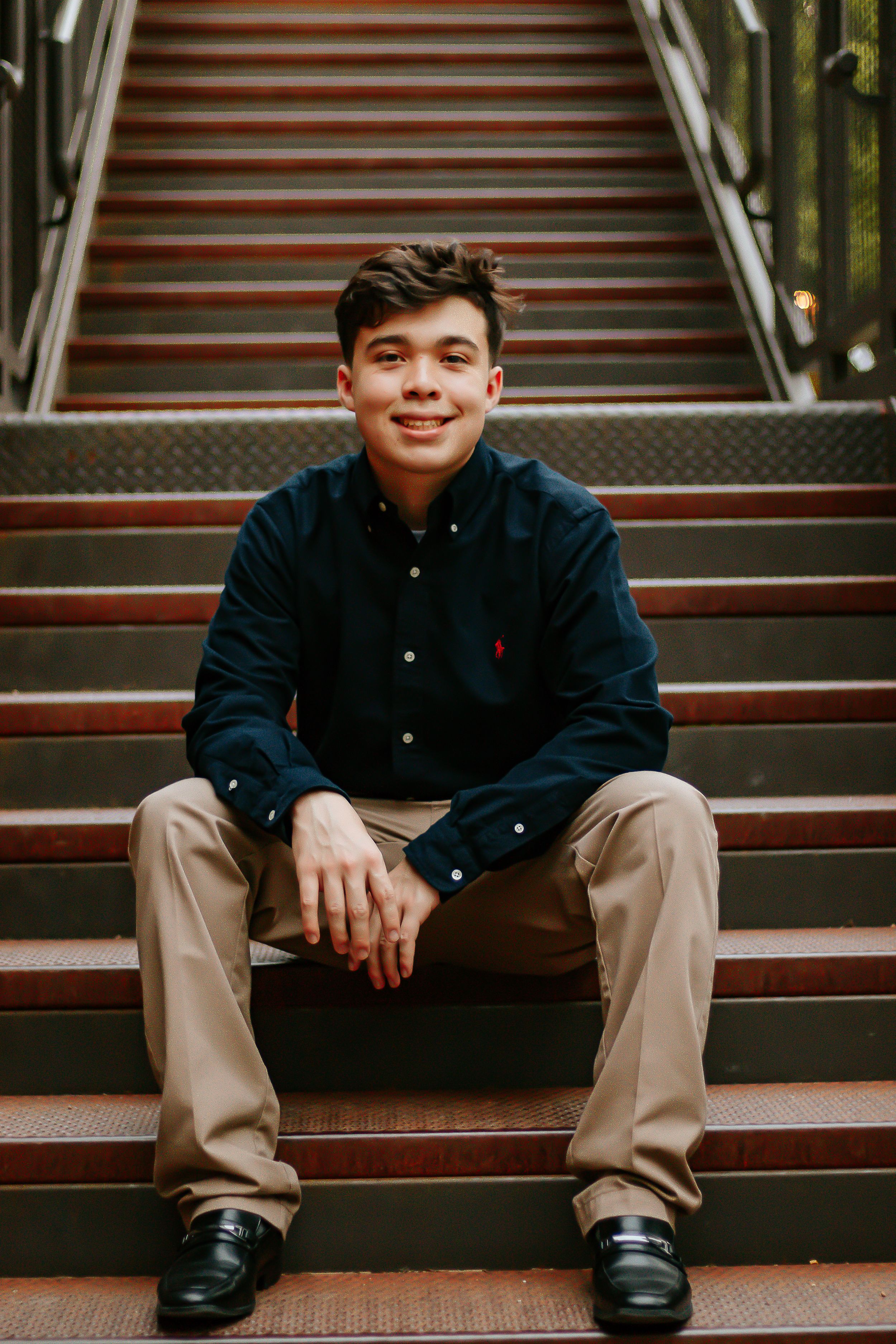 Young man in navy Polo shirt and khakis sitting on outdoor stairs, smiling confidently at camera.