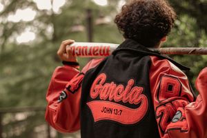 Person wearing a black and red Garcia varsity jacket with John 13:7 text, holding a baseball bat outdoors.