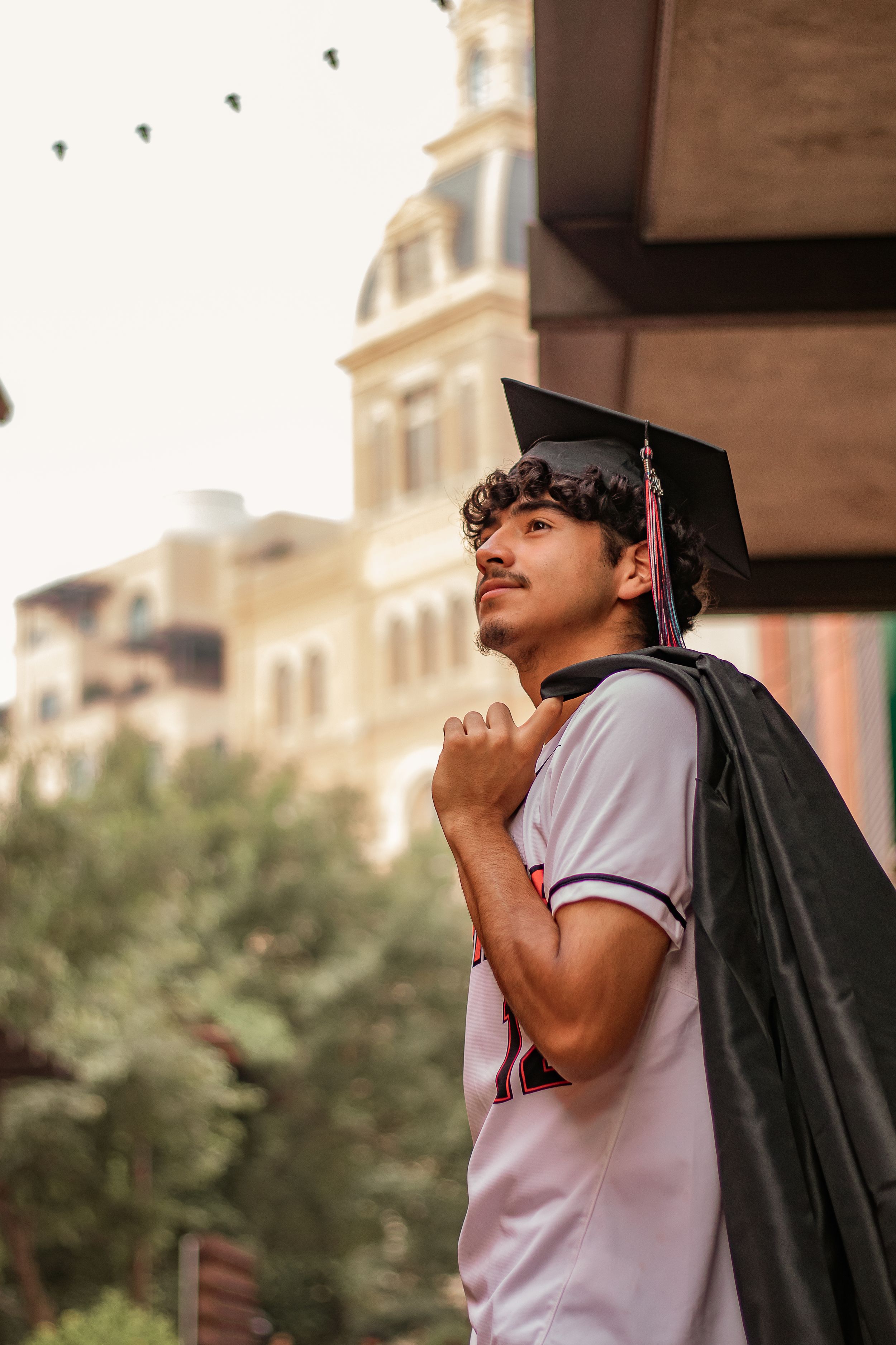 Young male graduate wearing cap and gown poses confidently outdoors near historic building with trees in background.