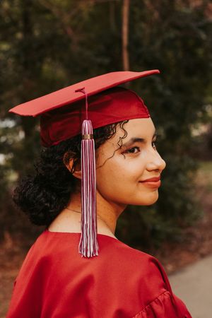 Young woman in red graduation cap and gown smiling outdoors, celebrating academic achievement.