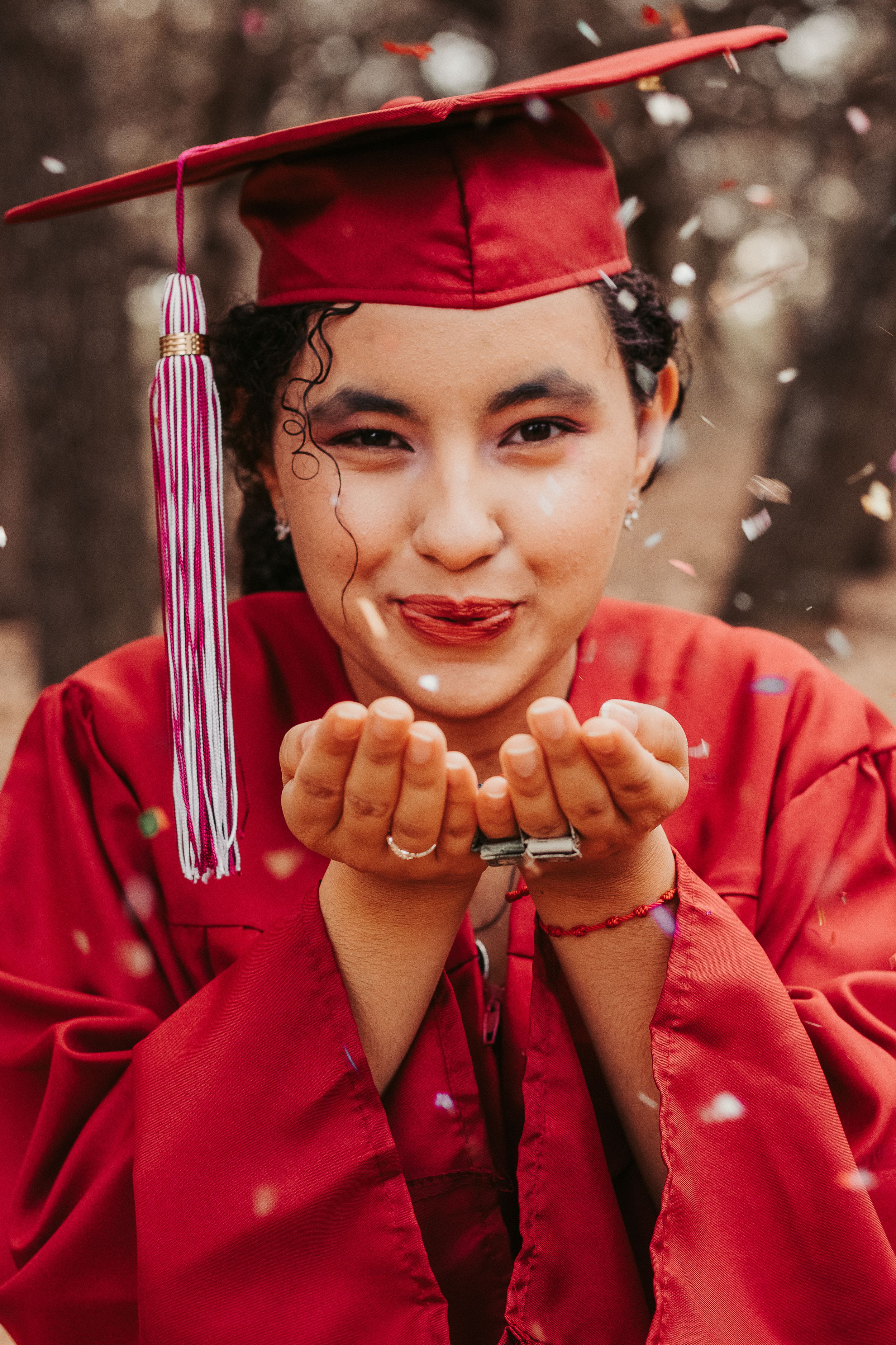Young woman in red graduation cap and gown blowing confetti from her hands, celebrating her achievement outdoors.