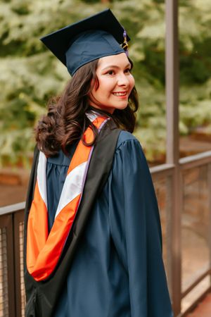Smiling young woman in navy blue graduation cap and gown with orange sash, posing outdoors near greenery.