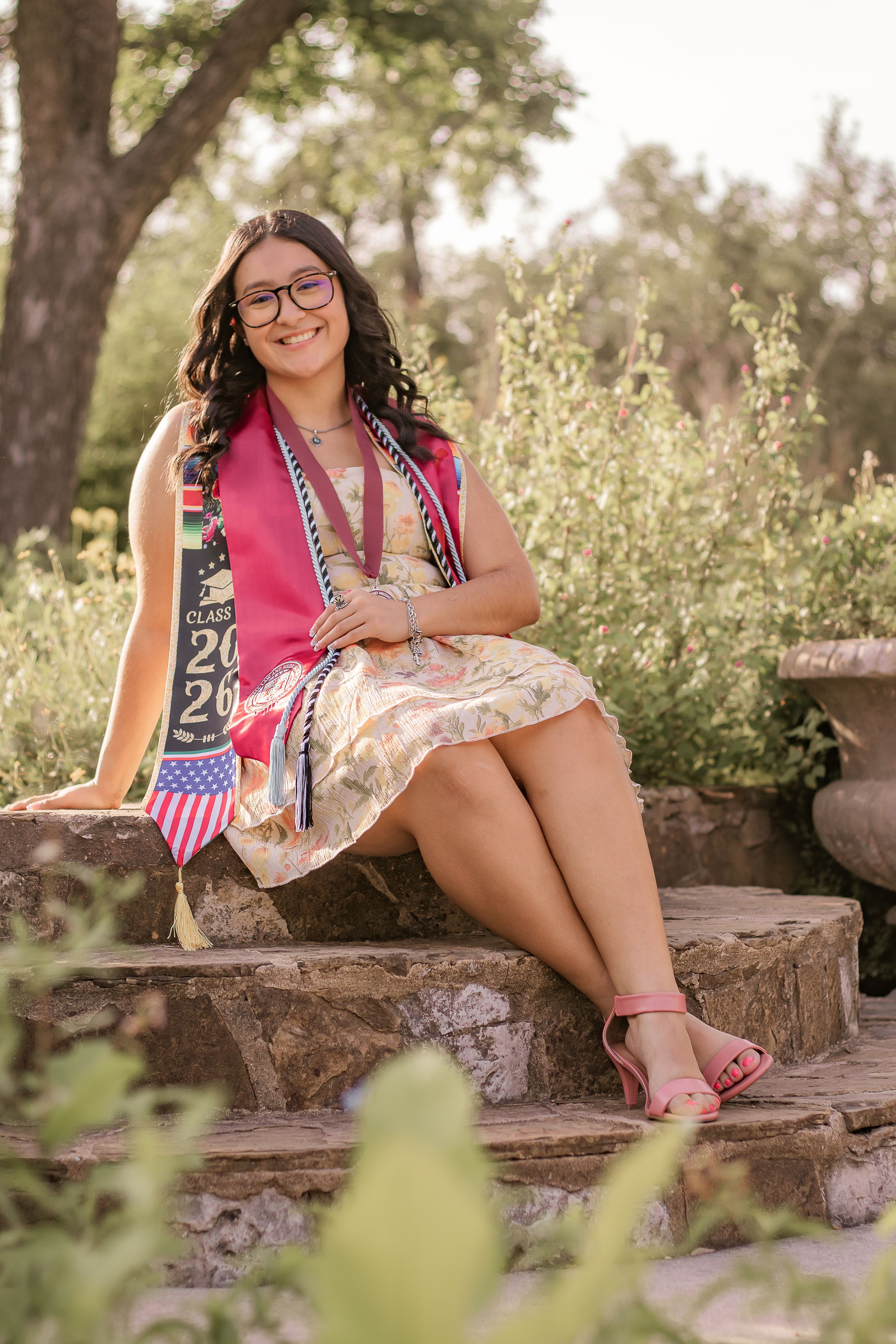 Smiling graduate sits on stone steps wearing a Class of 2026 sash and floral dress in a lush garden setting.