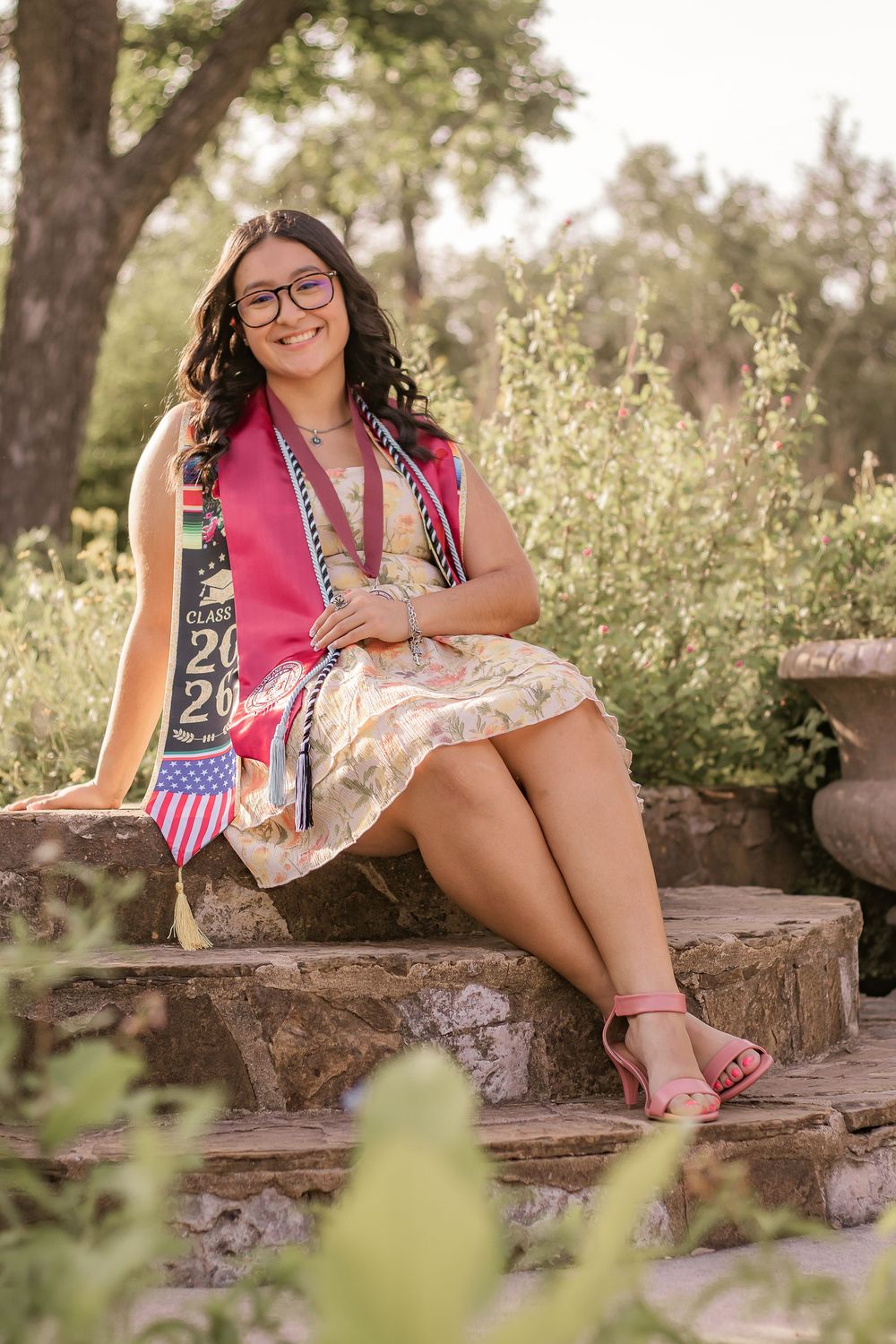 Smiling graduate sits on stone steps wearing a Class of 2026 sash and floral dress in a lush garden setting.