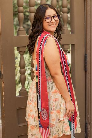 Graduate in floral dress with red sash and honor cords smiles over shoulder near ornate wooden doors.