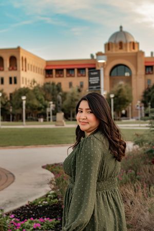 Young woman in green polka dot dress smiling over shoulder in front of a university building campus.