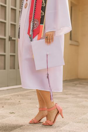 Graduate in white gown holding cap with pink tassel, wearing coral heeled sandals, adorned with red honor cords.