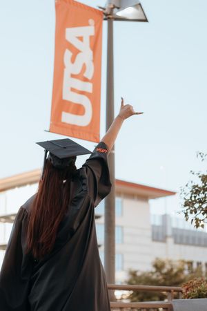 A UTSA graduate in black cap and gown raises hand toward orange UTSA banner on campus.