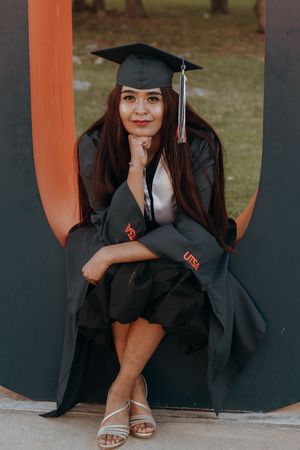 Young woman in black UTSA graduation cap and gown sitting proudly for a graduation portrait outdoors.