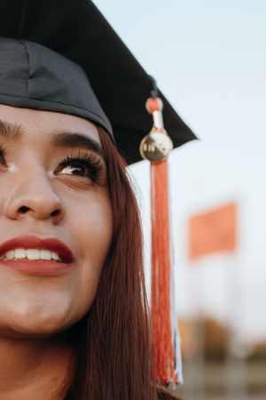 Close-up of a young woman in a black graduation cap with orange tassel, smiling and looking upward on graduation day.