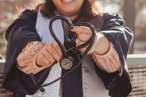 Woman holding a black stethoscope shaped like a heart, wearing a jacket outdoors.