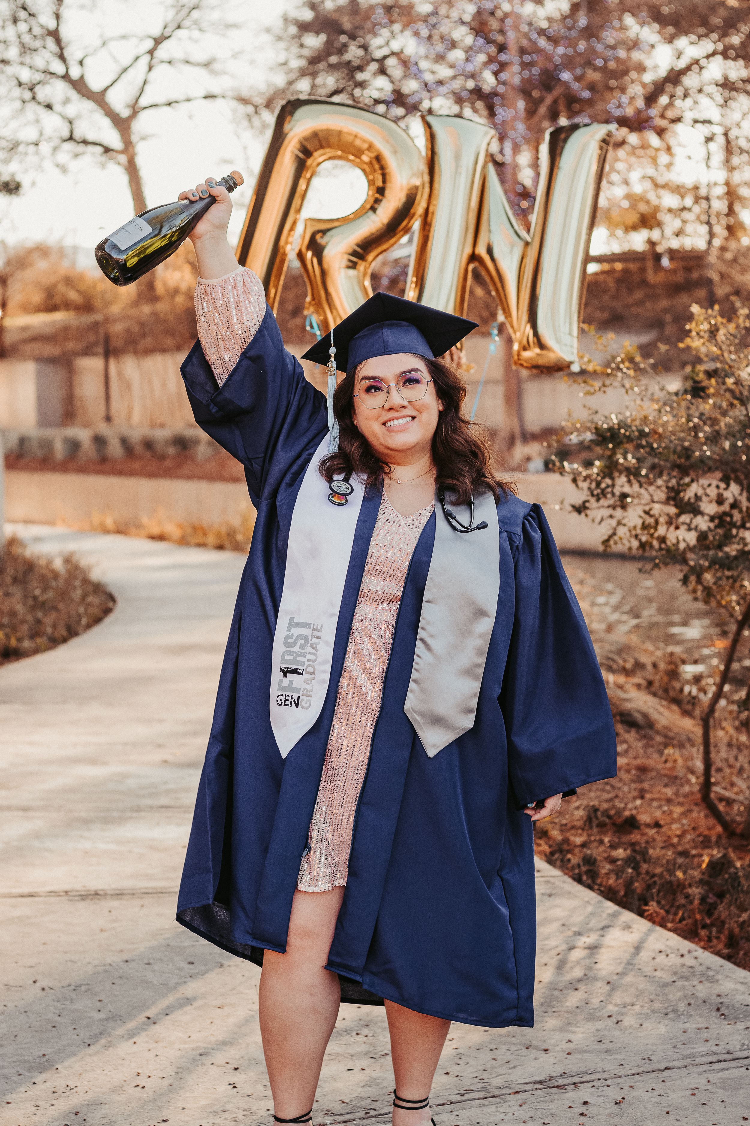 Nursing graduate celebrates in blue cap and gown, holding champagne and RN balloons outdoors.