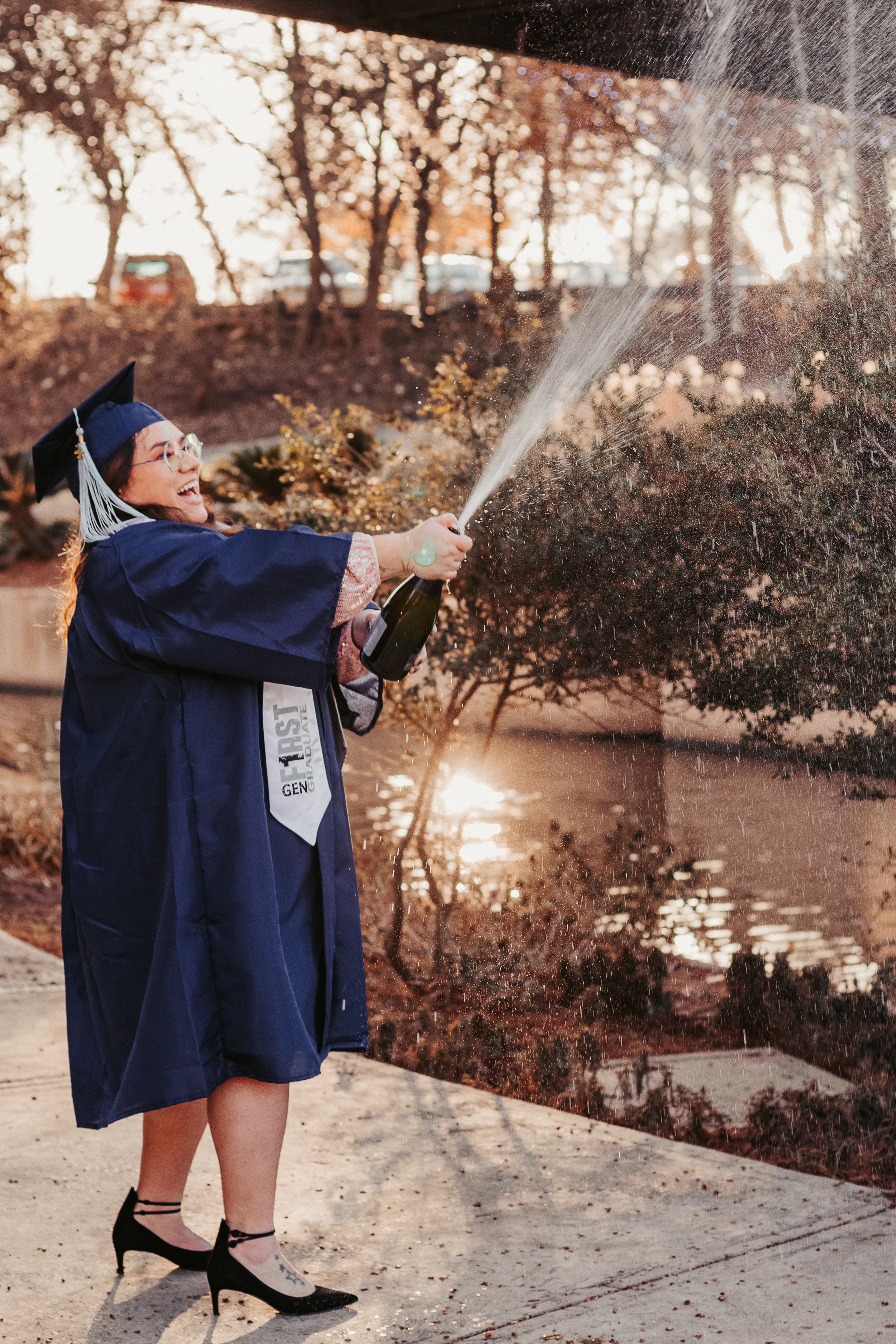 Graduate in navy cap and gown sprays champagne bottle outdoors, celebrating graduation milestone at sunset.