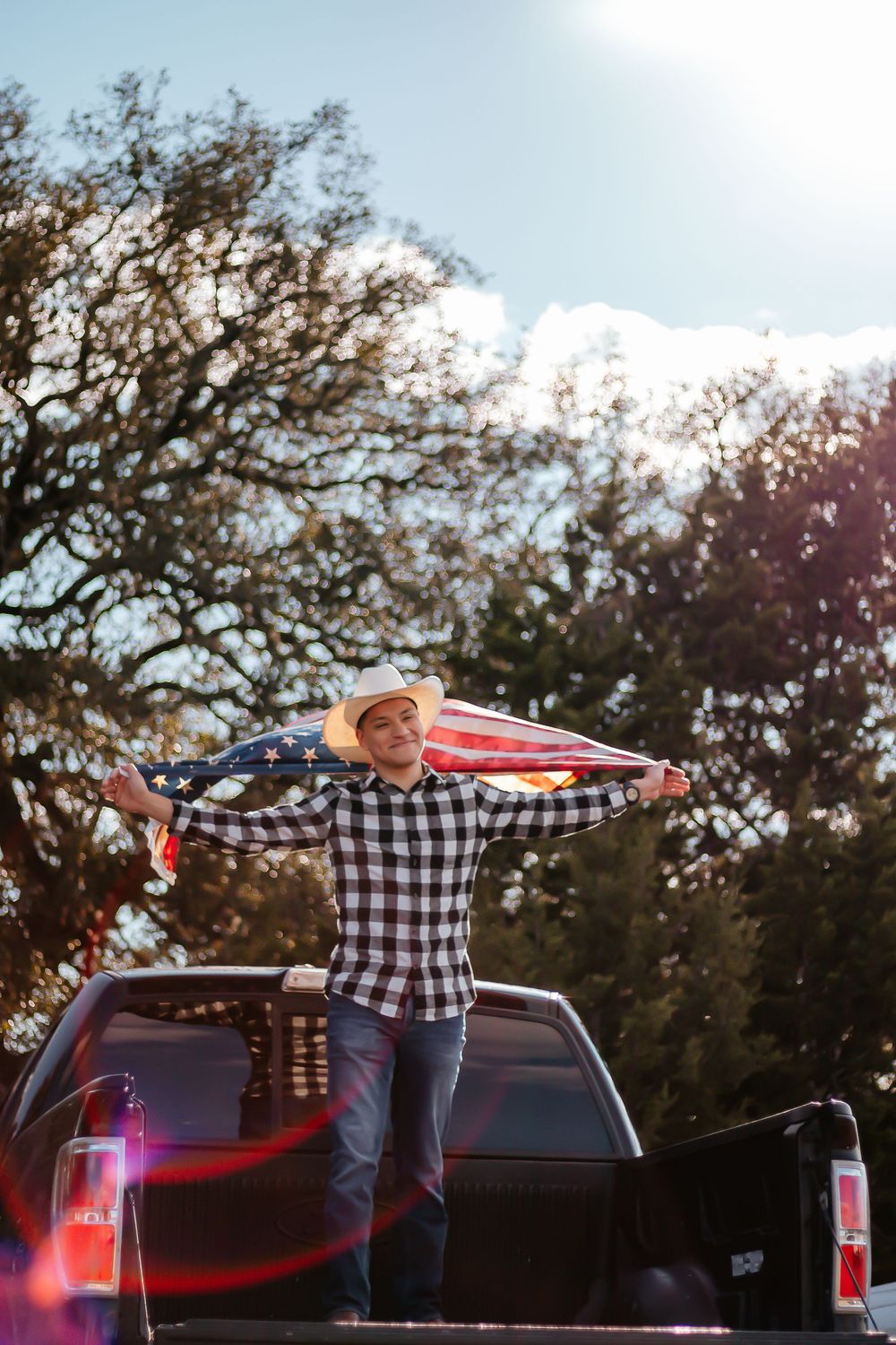 Young man in cowboy hat holding American flag while standing in truck bed outdoors on sunny day.