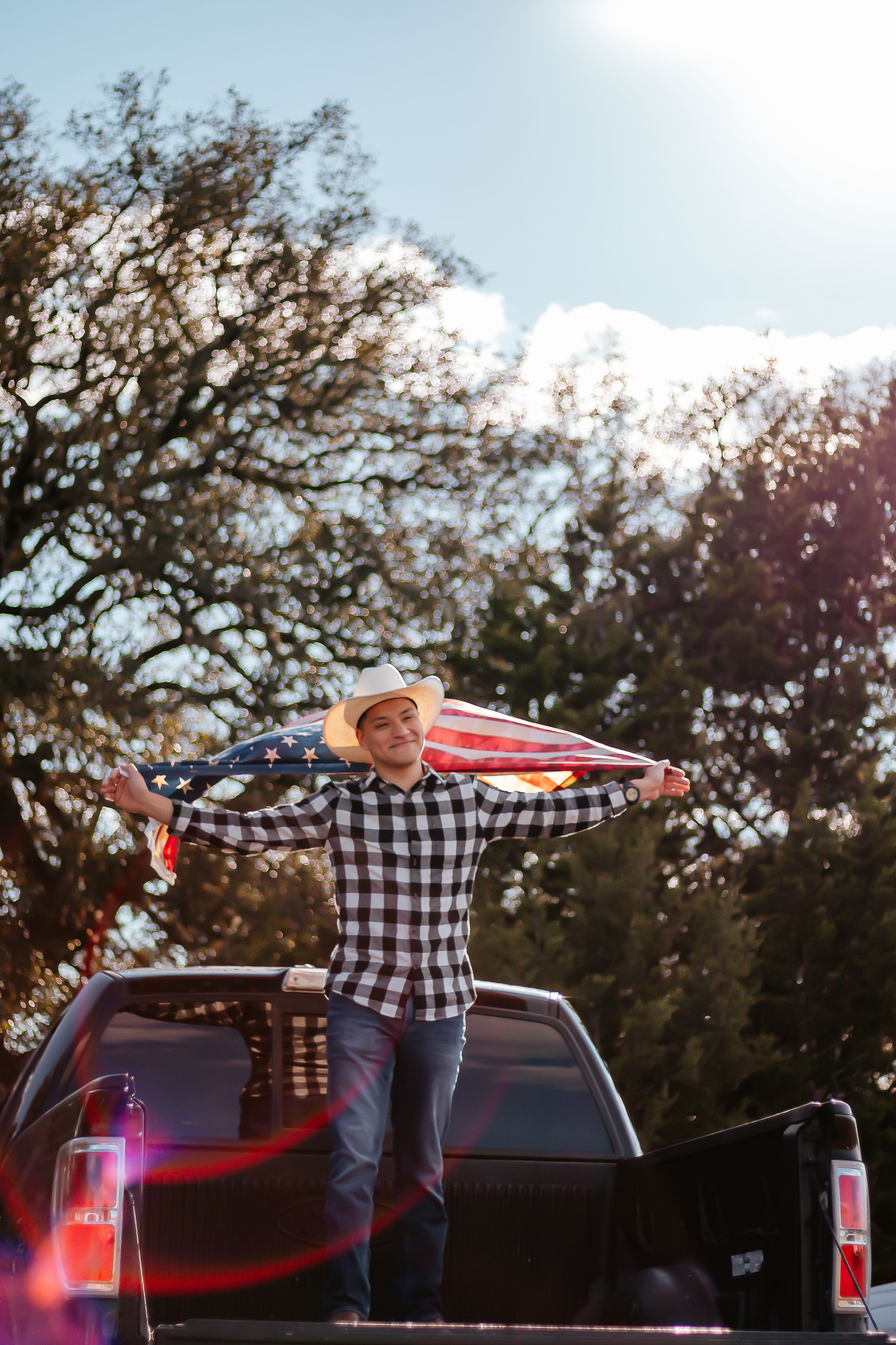 Young man in cowboy hat holding American flag while standing in truck bed outdoors on sunny day.