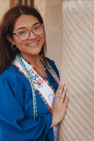 Smiling young woman in blue graduation gown with embroidered sash and glasses leaning against a stone column.