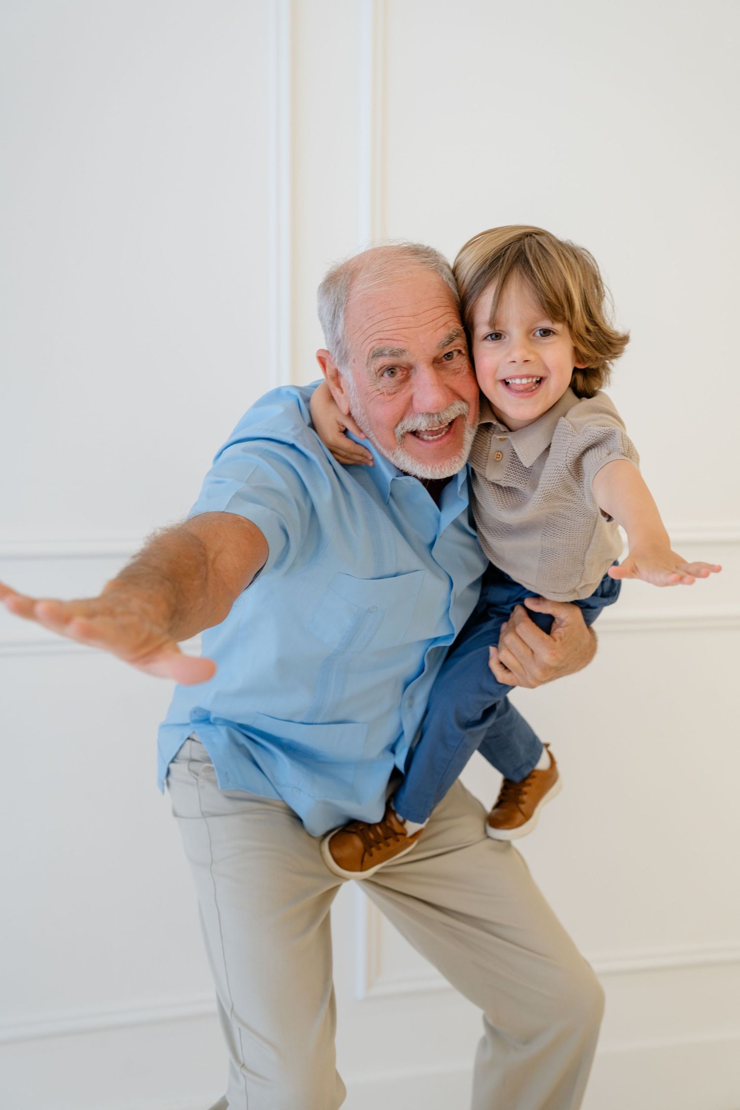 Elderly grandfather playfully holds young grandson with arms spread wide, both smiling joyfully in bright white room.