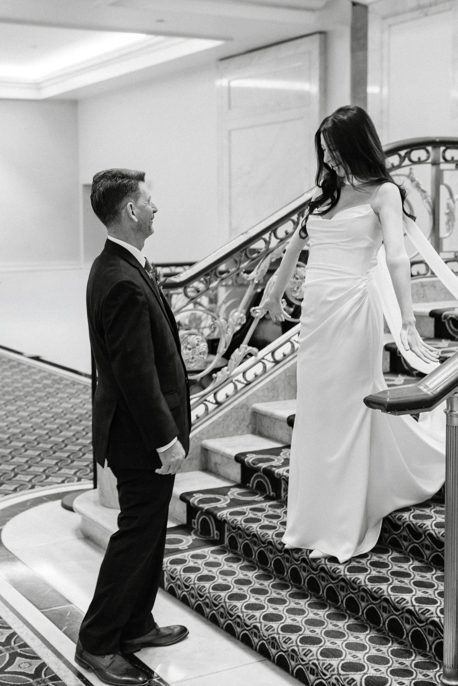 Black and white photo of bride in white gown and groom in tuxedo sharing a romantic moment on an elegant staircase.