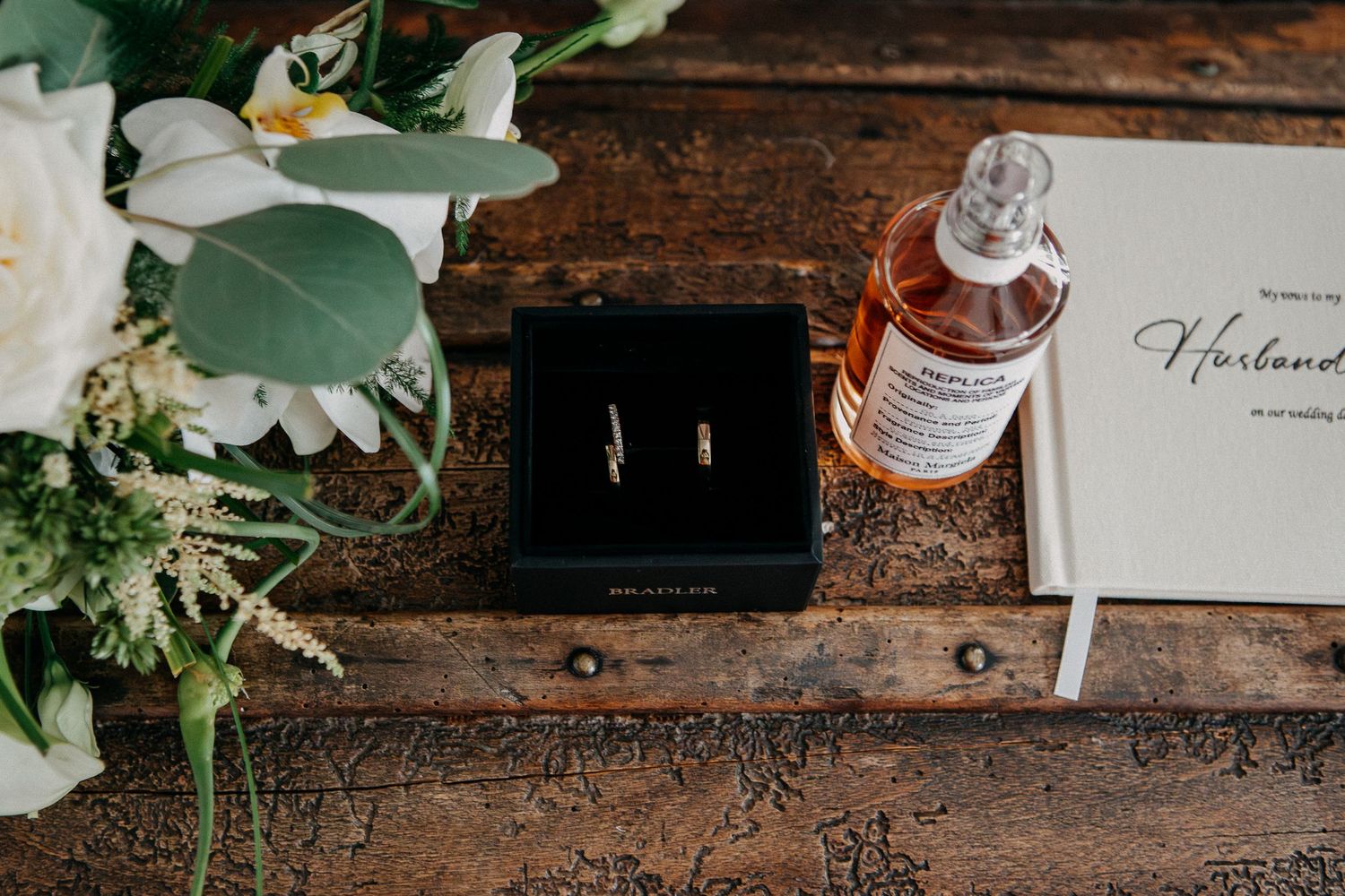 Wedding rings in black box beside whiskey bottle, bridal bouquet, and Husband vow book on rustic wood table.