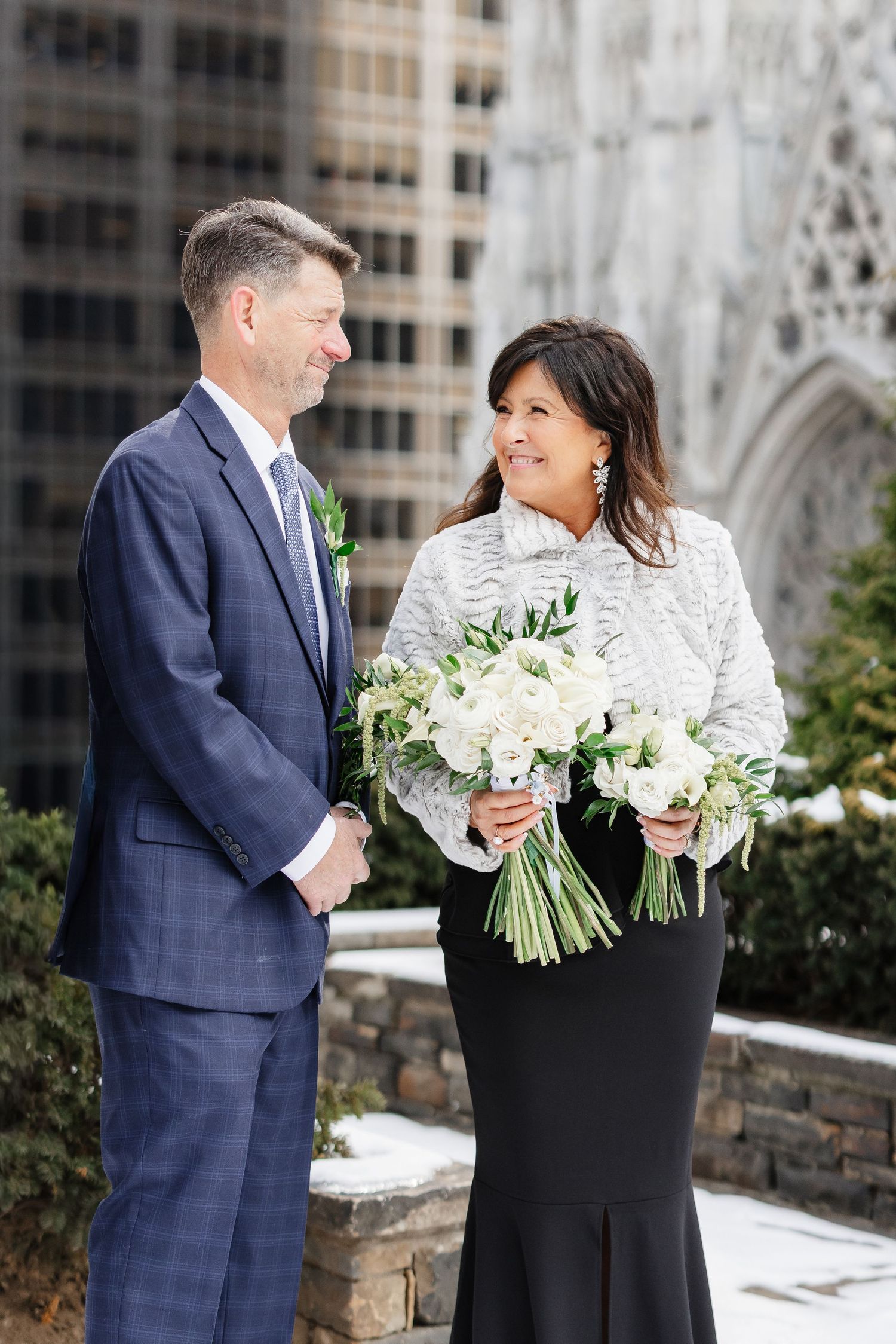 Couple in formal attire smiling at each other outdoors, bride holding white floral bouquet with city architecture background.