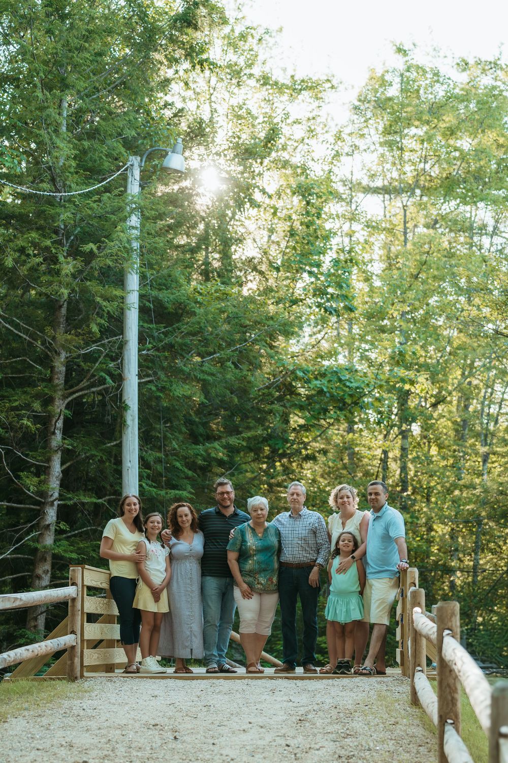 Multi-generational family posing together on a wooden bridge surrounded by lush green trees on a sunny day.