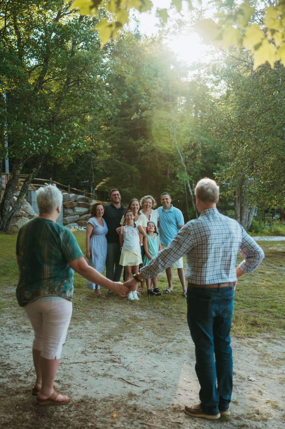 An elderly couple holds hands while facing their multigenerational family gathered outdoors among lush green trees.