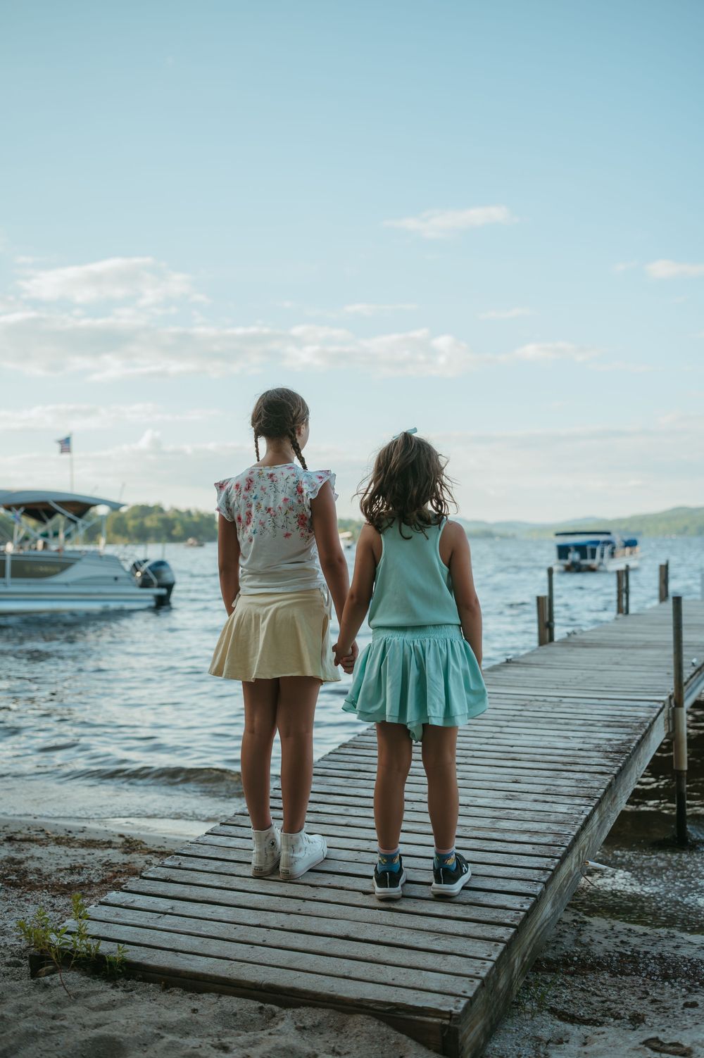 Two young girls holding hands stand on a wooden dock overlooking a calm lake with boats in the distance.