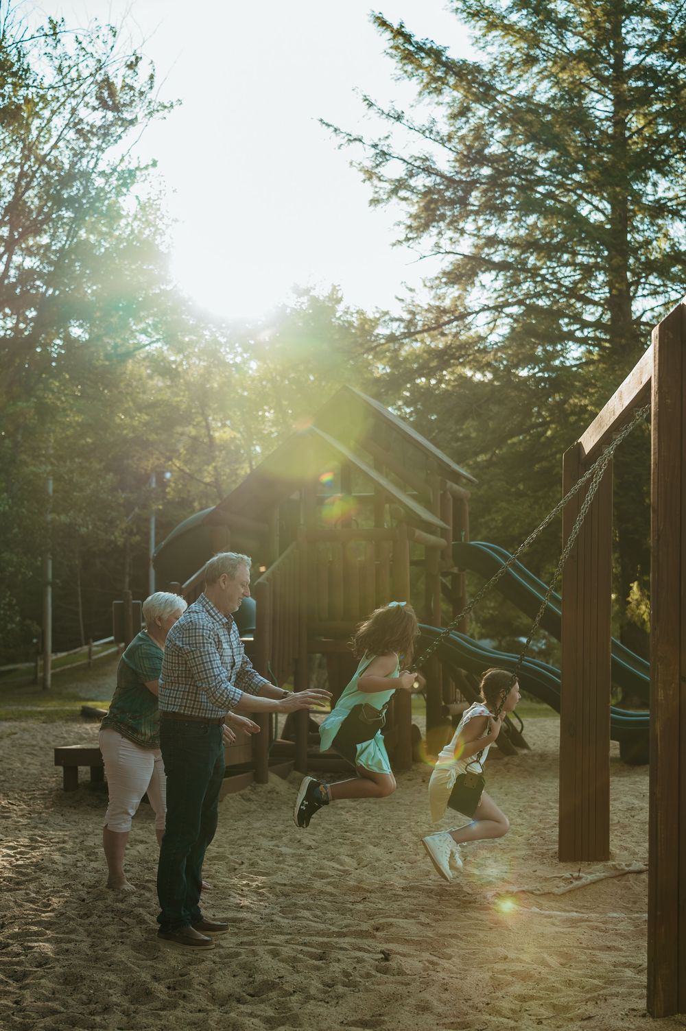 Family enjoying a sunny playground with children on swings as an adult pushes them in a forest setting.