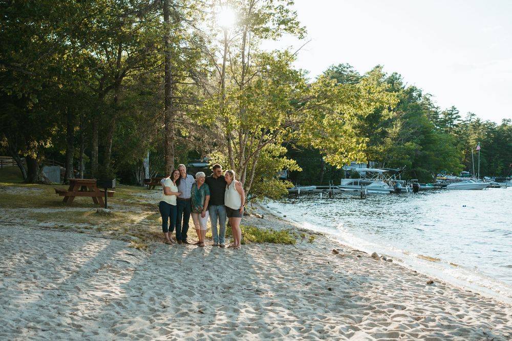 Group of people posing together on a sandy beach shore surrounded by lush green trees in warm sunlight.