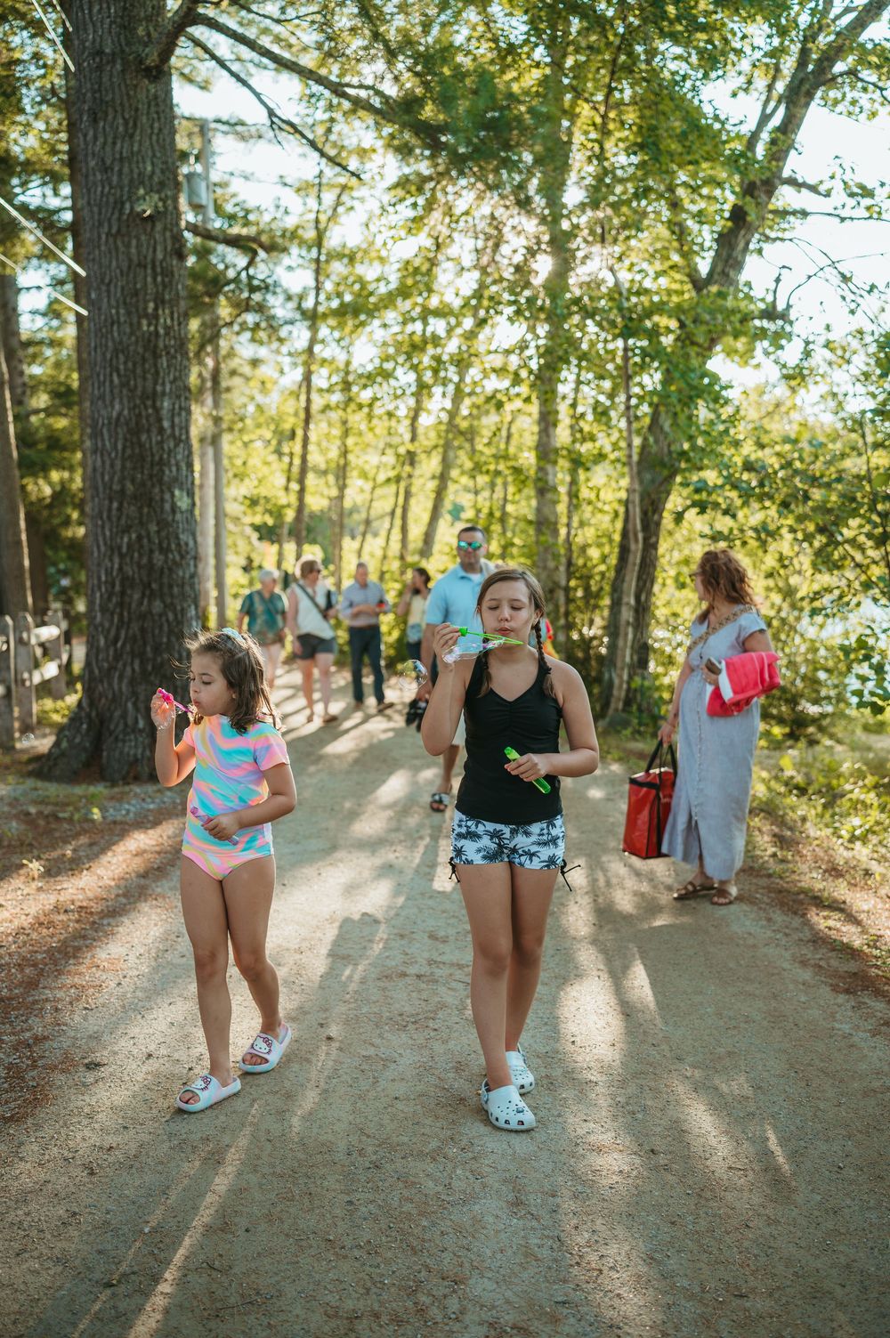 Two girls blowing bubbles on a sunny wooded path with adults in the background on a warm summer day.