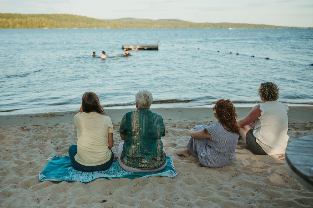 Four people sitting on a blanket on a sandy beach, watching swimmers in calm blue water at dusk.