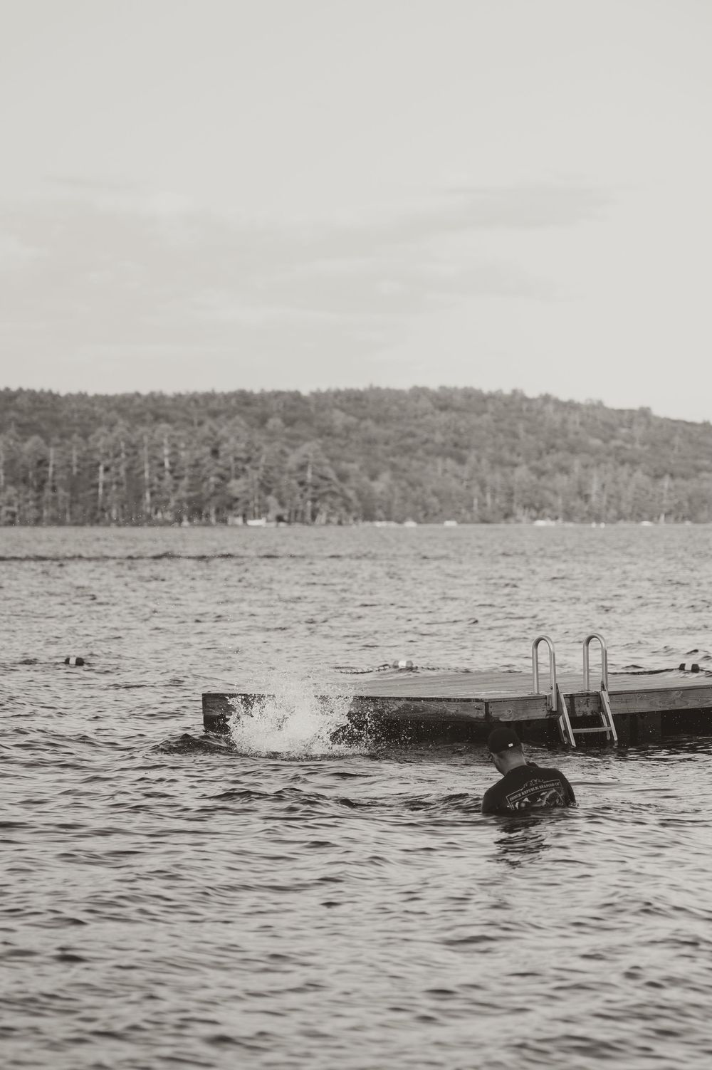 Black and white photo of a person jumping off a lake dock with splashing water and forested hills in the background.