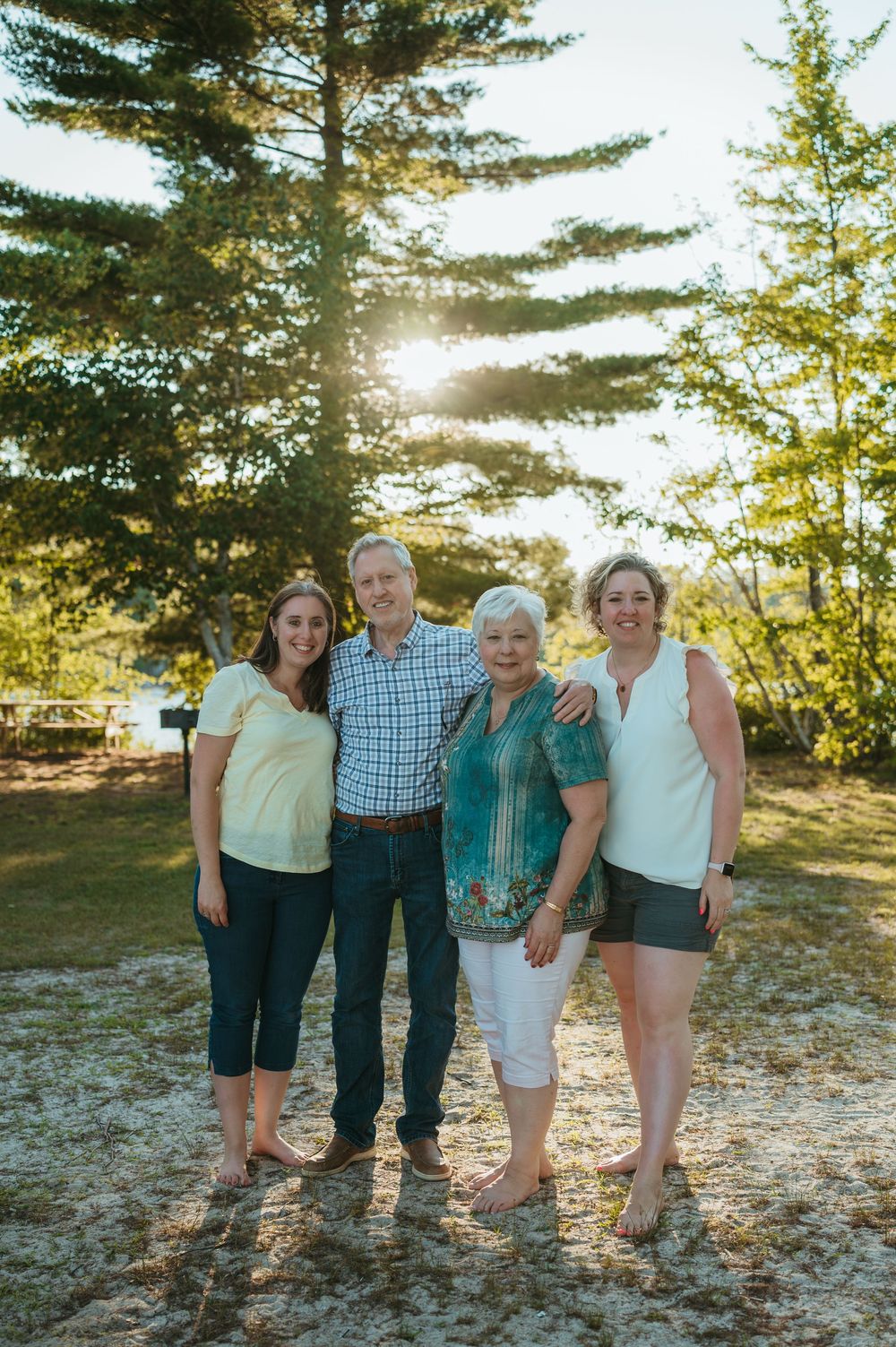 Family of four smiling outdoors near tall pine trees with sunlight filtering through on a warm summer day.