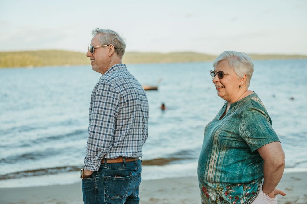 Elderly couple standing on a sandy beach by calm blue water, wearing sunglasses and casual clothing on a cloudy day.