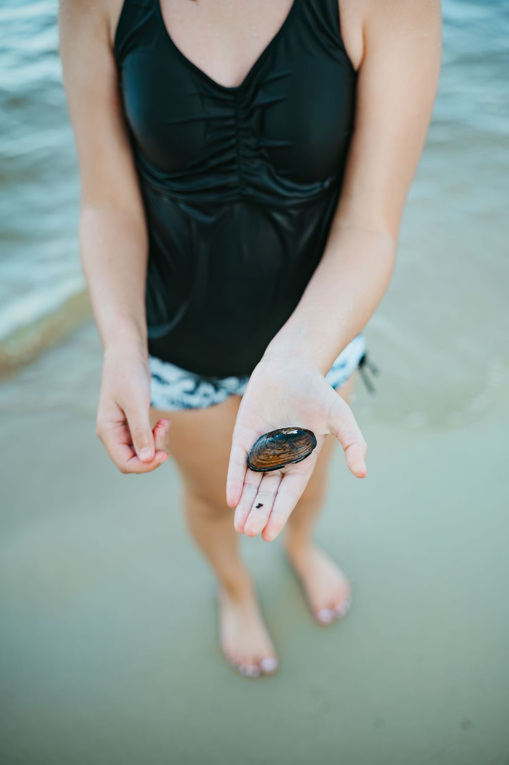 Woman in black swimsuit holding a dark shell on her palm while standing in shallow ocean water.