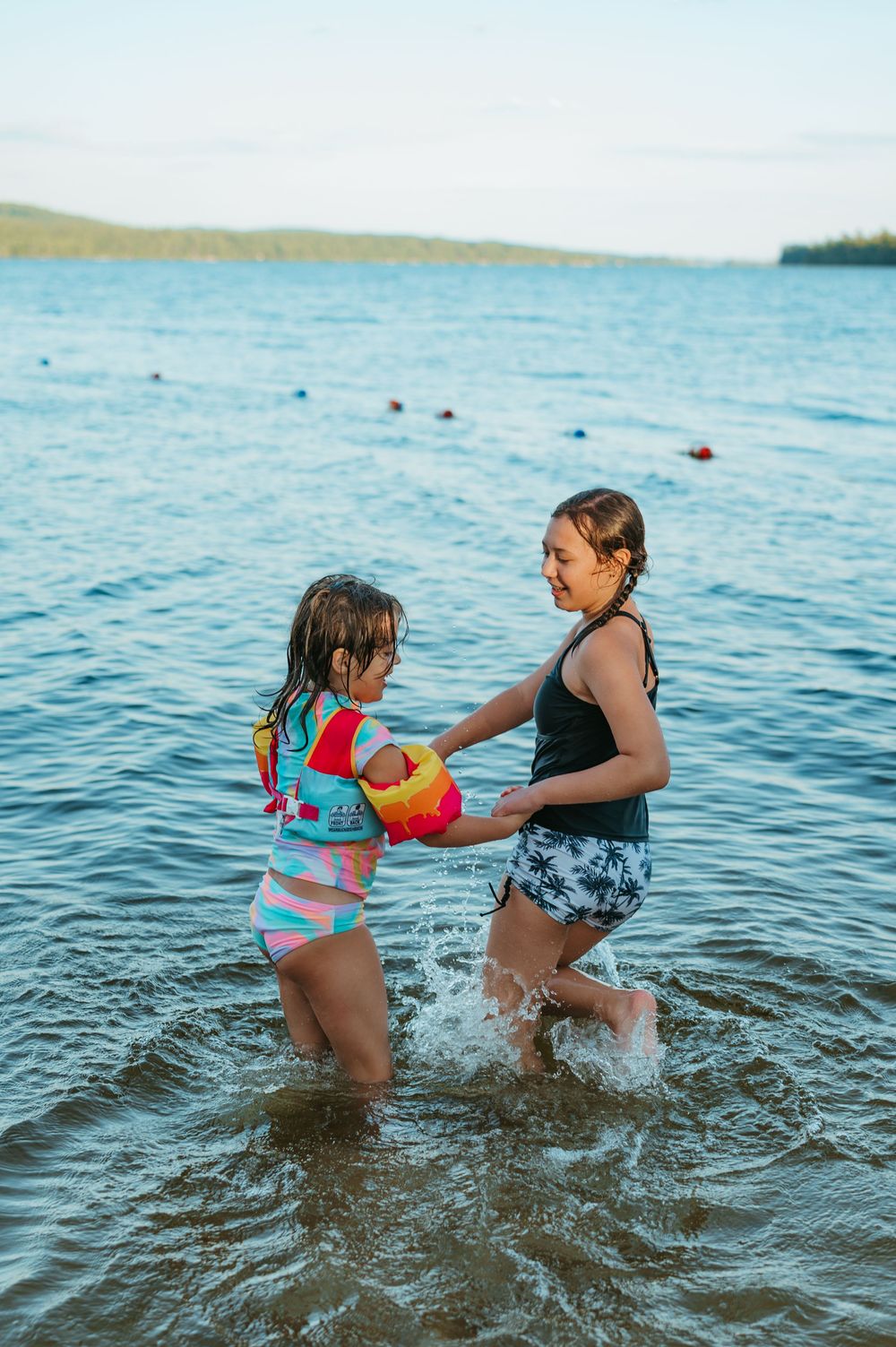 A young girl in a colorful life jacket splashes in a lake with an older girl on a sunny evening.