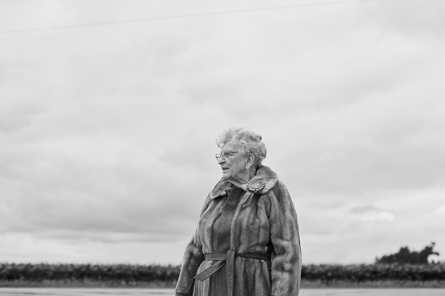 Elderly woman in fur coat standing outdoors under cloudy sky, black and white portrait photography.