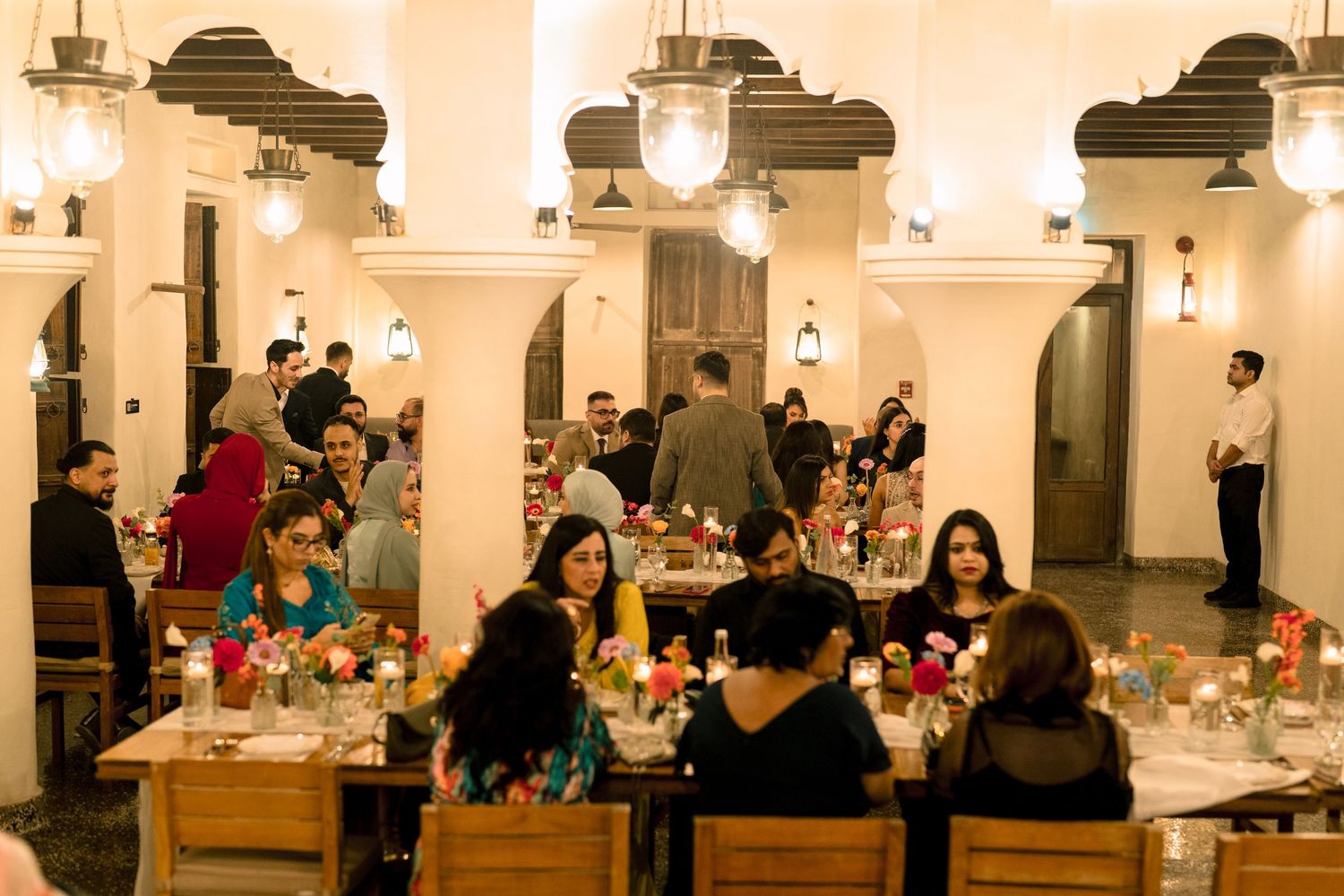 Guests dining at elegant restaurant with warm lighting, colorful floral centerpieces, and arched white walls.