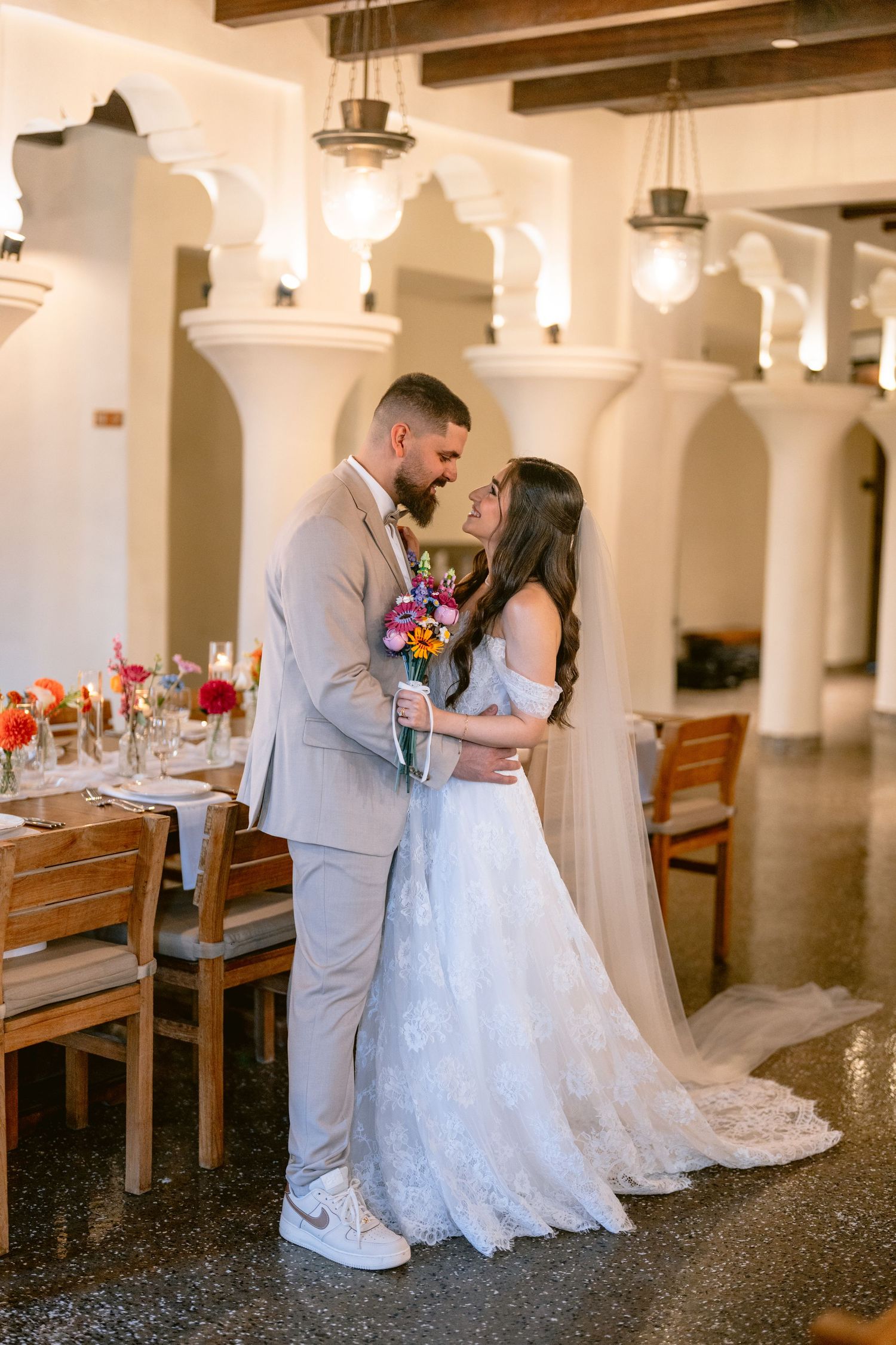 Bride and groom share a romantic moment in elegant venue, bride in lace gown with veil holding colorful bouquet.