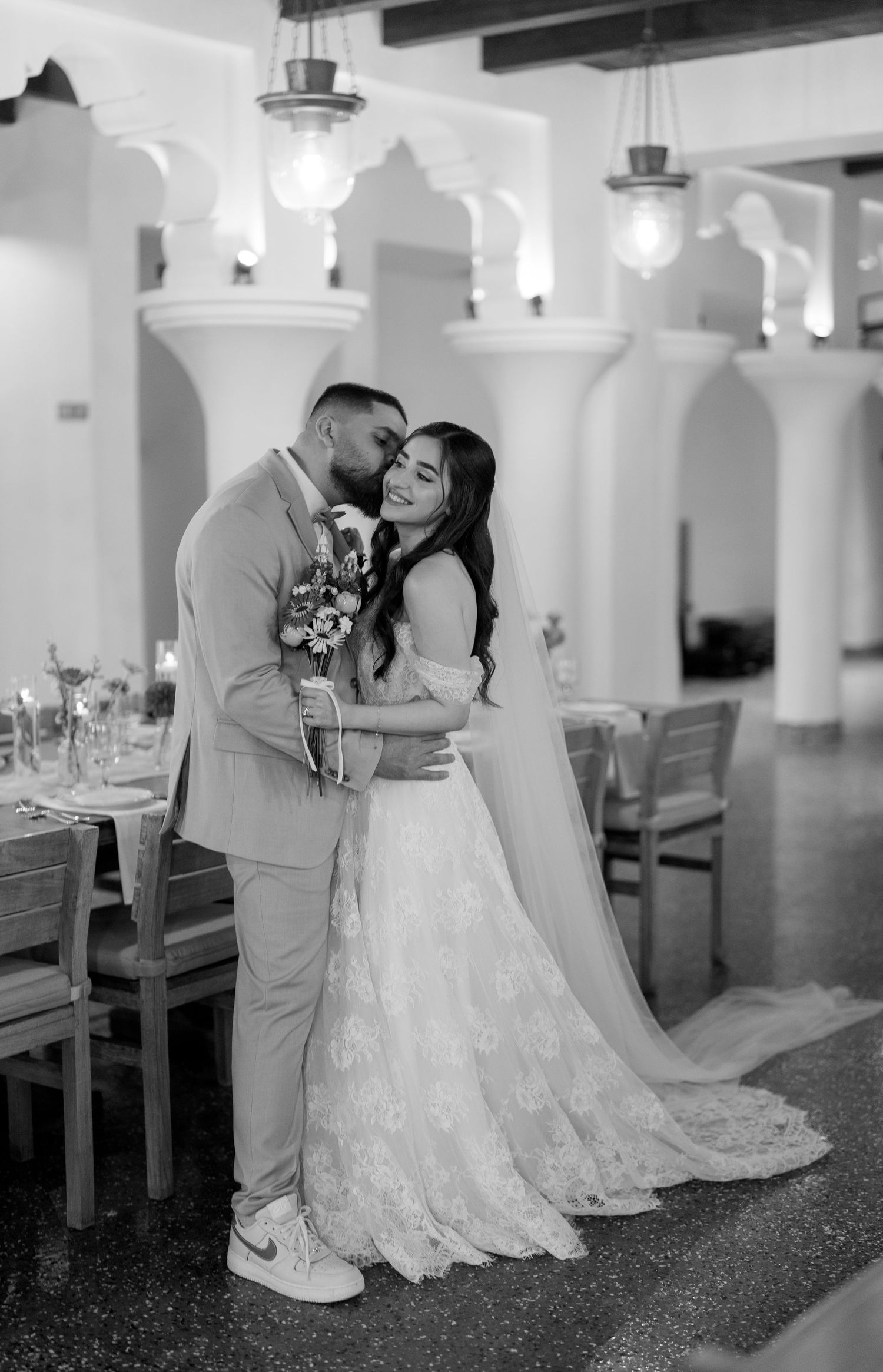 Black and white wedding photo of bride in lace gown and groom in suit sharing a romantic embrace in elegant venue.