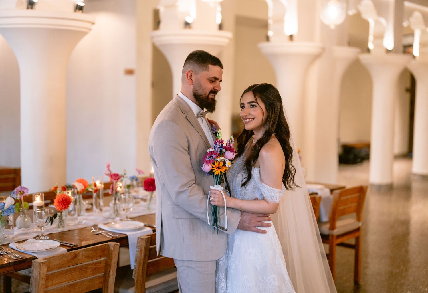 A bride and groom share a romantic moment in an elegant venue with colorful floral arrangements and arched columns.