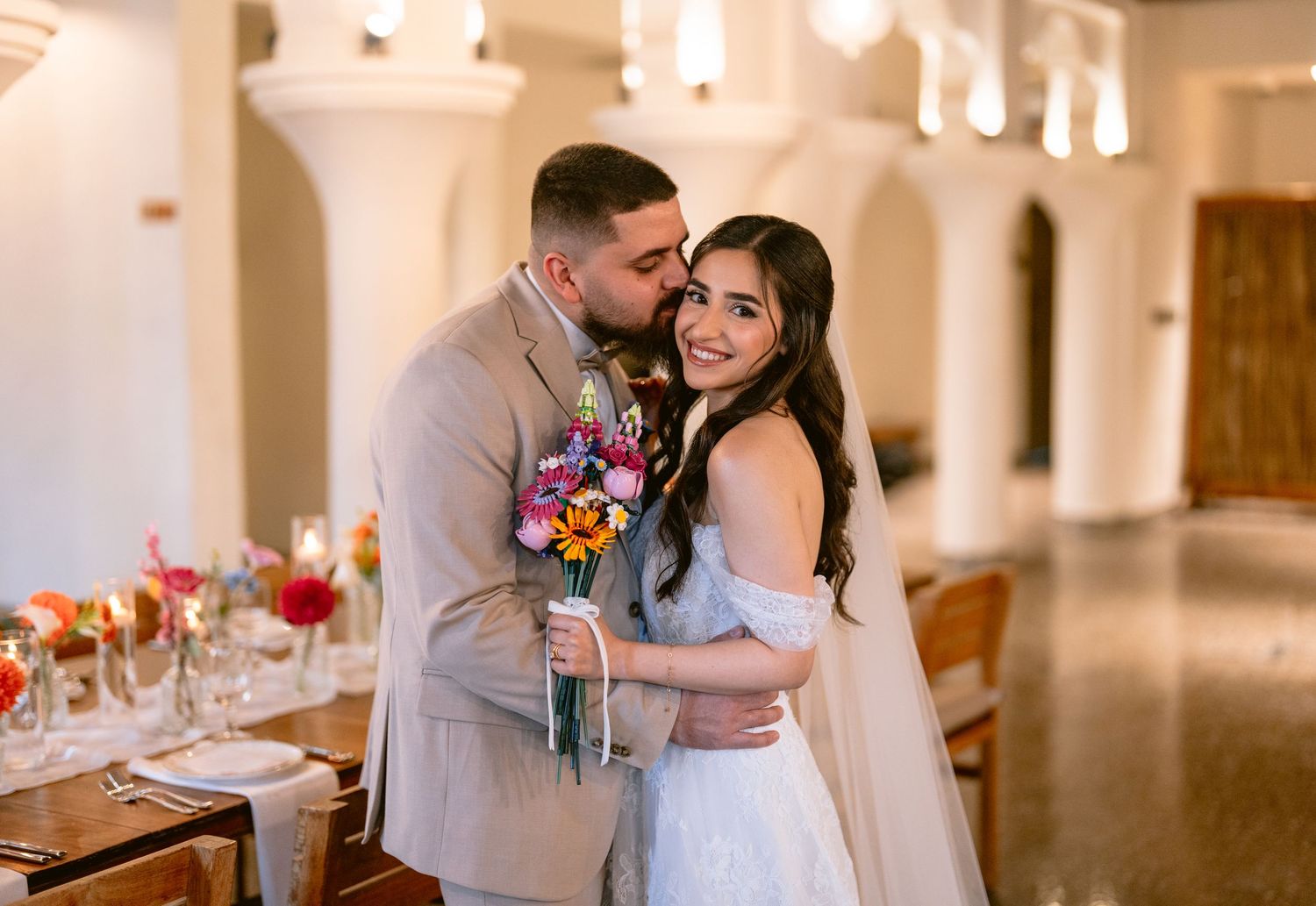 Groom in tan suit kisses bride in white gown holding colorful bouquet inside elegant wedding venue.