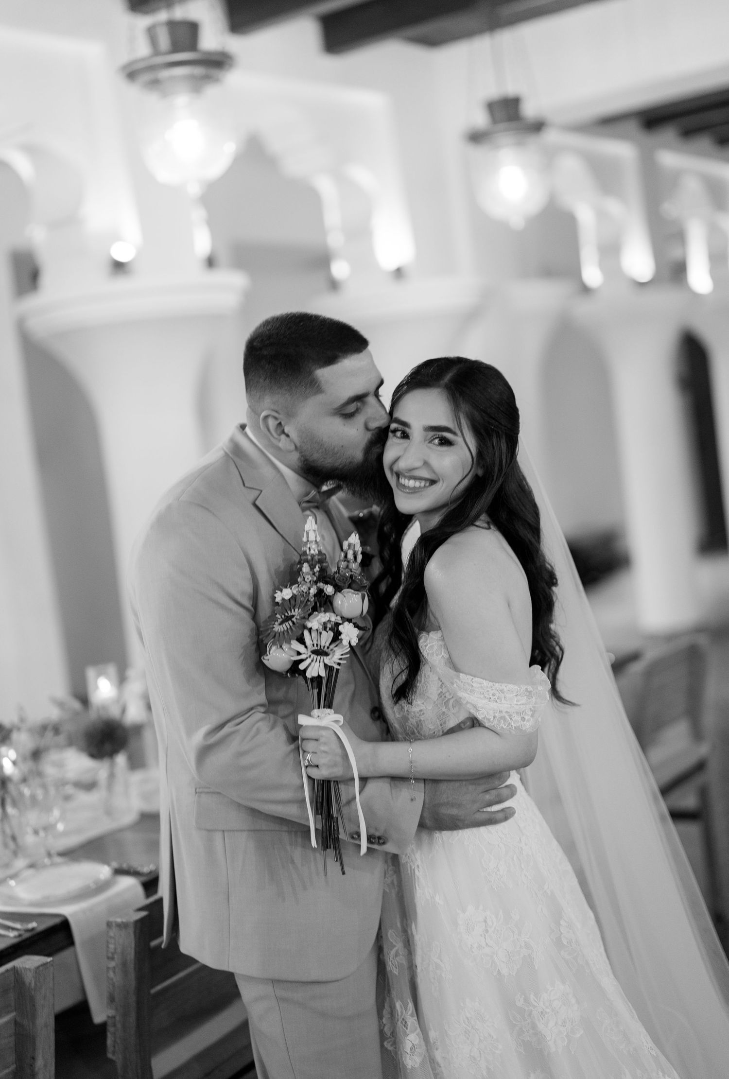 Black and white wedding photo of groom kissing bride on cheek while she smiles holding a bouquet in elegant venue.
