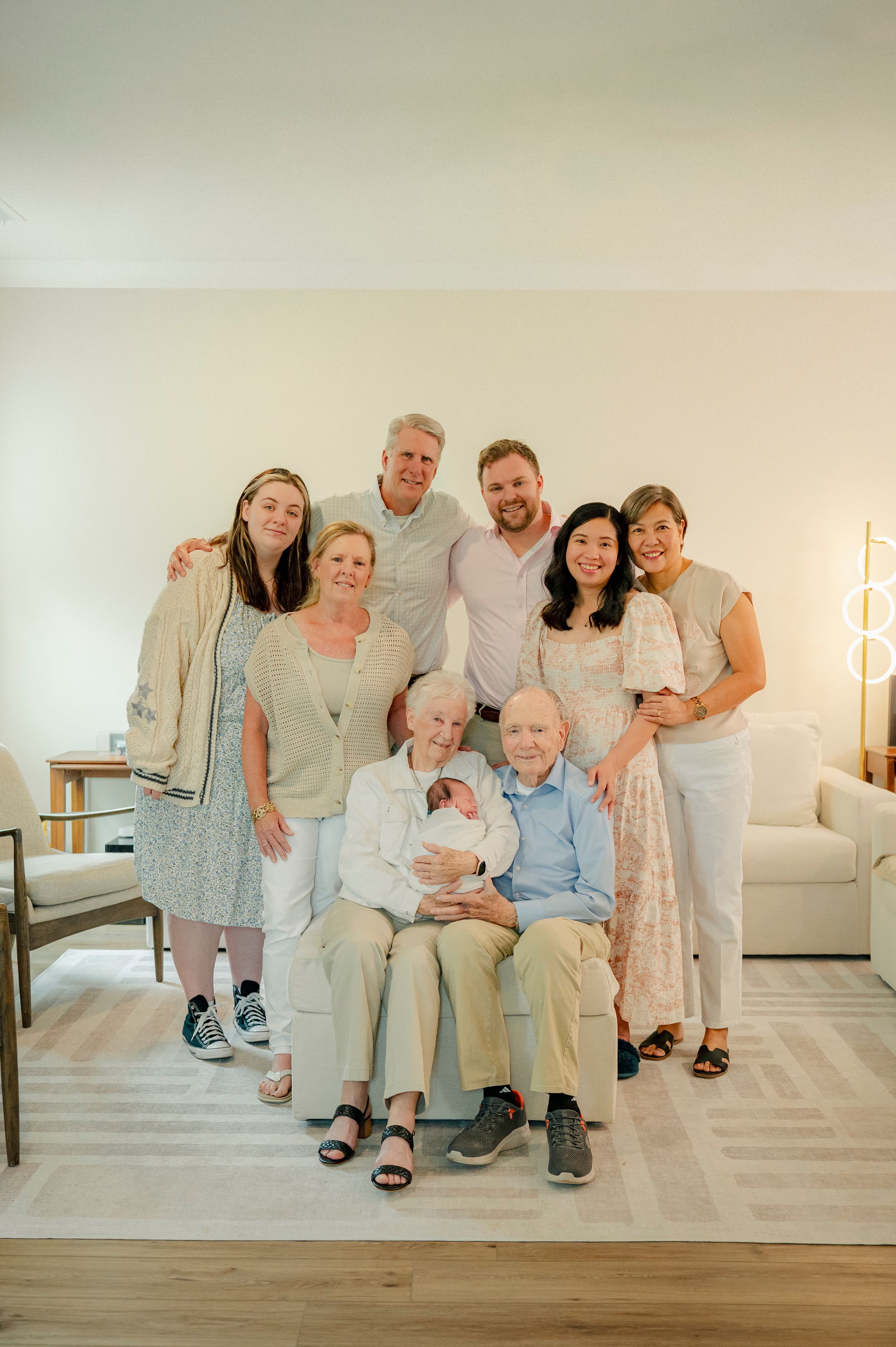Multi-generational family portrait with grandparents seated holding newborn baby, surrounded by smiling relatives in bright living room.