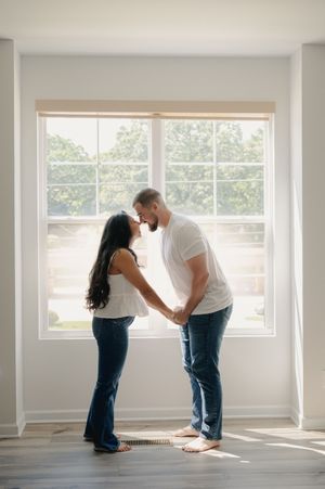 A couple shares a romantic moment holding hands and almost kissing in front of a bright window in an empty room.