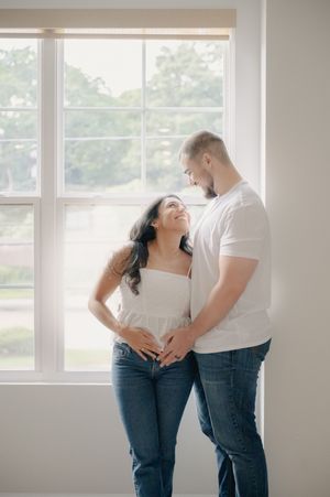 A couple smiles at each other while standing by a bright window, wearing white tops and jeans in a cozy indoor setting.