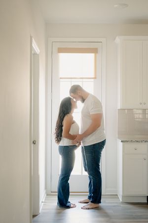 A couple shares a tender kiss while standing in a sunlit kitchen doorway, both wearing white tops and jeans.
