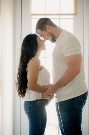 Couple sharing a tender moment near a bright window, woman showing a baby bump in a maternity photo shoot.