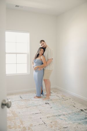 Pregnant woman in blue dress embraced by partner in striped shirt, posing in bright minimalist room.