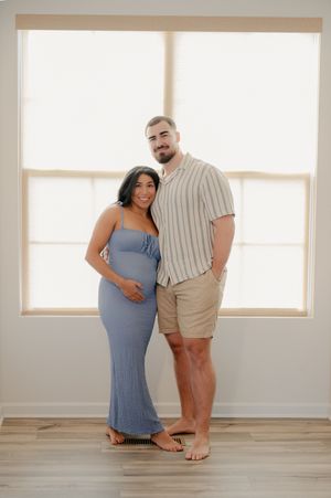 Smiling pregnant couple posing together in a bright room, woman in blue dress cradling baby bump, man in striped shirt.
